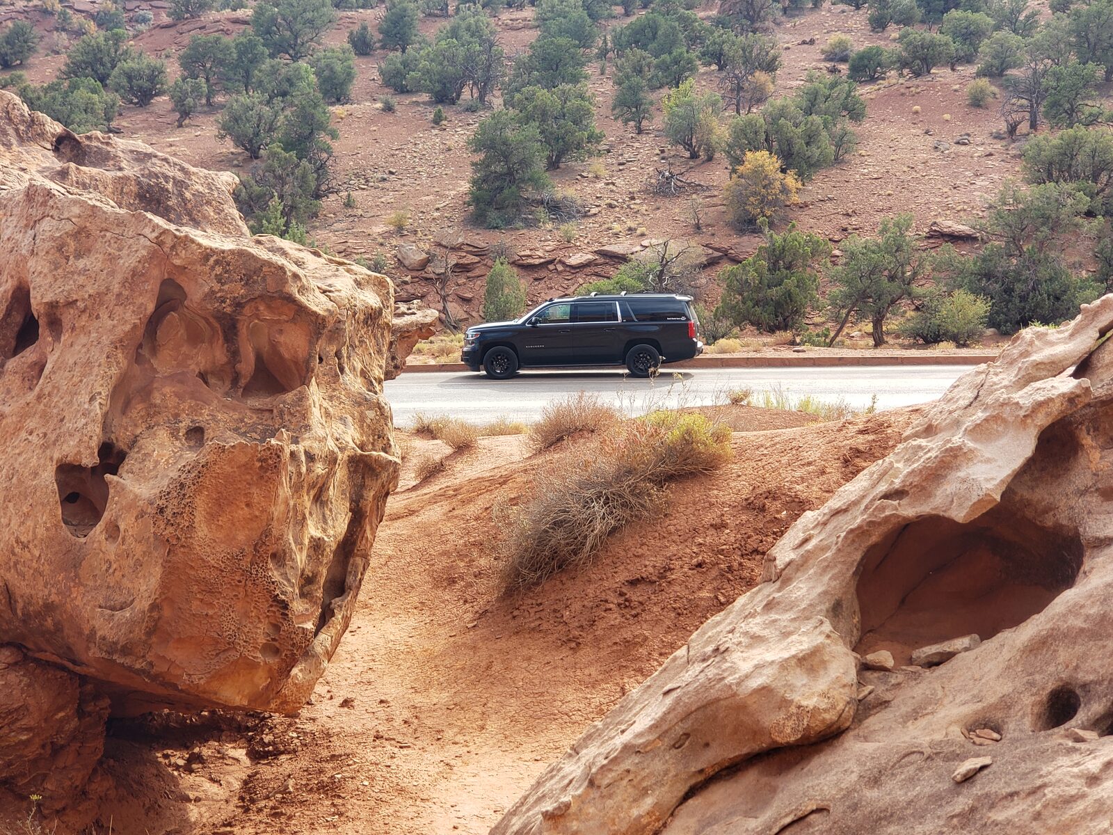 The full-size SUV on the shoulder of a two-lane road in southern Utah, framed between two weathered sandstone boulders